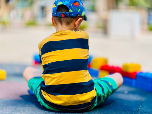 A young boy in a striped shirt and colorful cap sits on the playground, arranging large building blocks in various colors.
