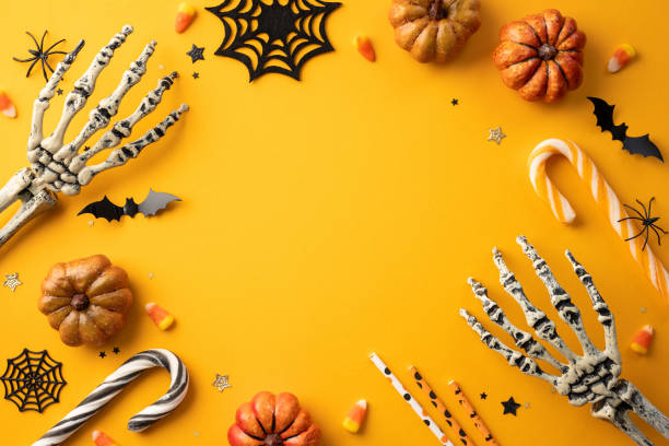 istock Festive Halloween background featuring skeleton hands, pumpkins, candy corn, spider webs, and bats on a yellow backdrop 2165250008