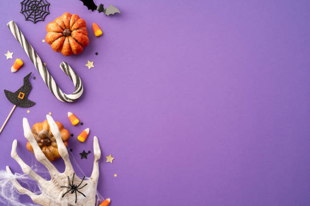 istock Purple background with Halloween decorations including pumpkins, candy corn, witch hat, and skeleton hand 2165250019
