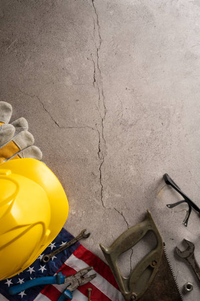 istock Yellow hard hat, work gloves, tools, and American flag on a cracked vertical concrete surface. Labor Day in USA concept 2165250033
