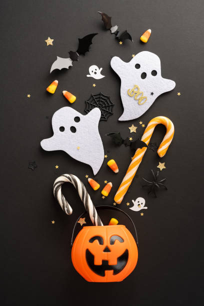 istock Festive vertical Halloween background with cute ghost figures, candy corn, peppermint sticks, bats, and a smiling pumpkin bucket on a black surface 2165250054