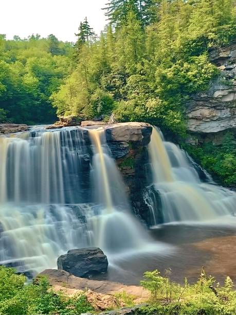 A long exposure photo of Blackwater Falls captures the ethereal beauty of the cascading waters as they flow over the rugged rock formations. The silky smooth effect of the water creates a stunning contrast against the sharp details of the surrounding landscape. Rich, dark tones of the blackwater add depth and mystery, while the blurred motion of the water provides a sense of tranquility and timelessness. The foliage and rocks around the falls are crisply detailed, emphasizing the serene yet powerful nature of this iconic West Virginia landmark. The overall composition highlights the dynamic interplay between motion and stillness, creating a mesmerizing and otherworldly scene.