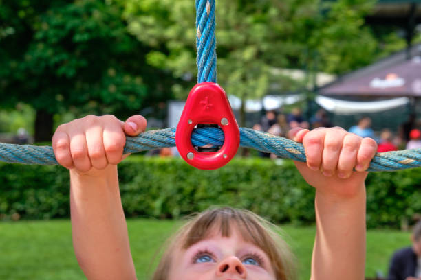A close-up of a young child as they reach up, grasping a thick blue rope on a playground. The child's hands tightly clutch the rope, and their eyes are focused upward with determination, as if gauging the next move. The vibrant red plastic connector on the rope contrasts with the greenery in the background, highlighting the intensity of the moment. This image beautifully captures the theme of challenge and perseverance during childhood play, emphasizing the child's focus and resolve in the midst of a playful yet determined effort.