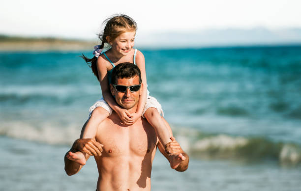 Father holding daughter at shoulders at sea coast portraits. Family dad and little pretty child kid at beach at summer together