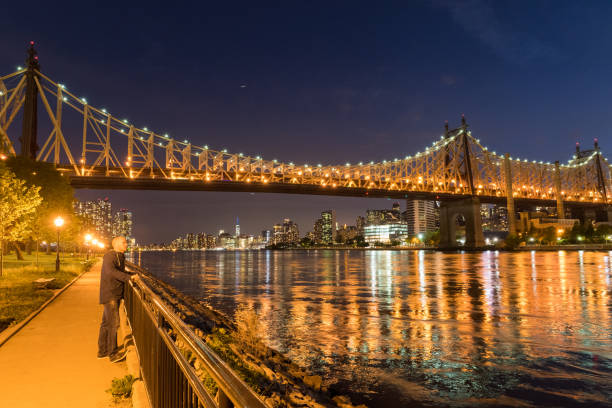 Man stands on riverbank below bridge at night, Brooklyn Bridge, Brooklyn, New York