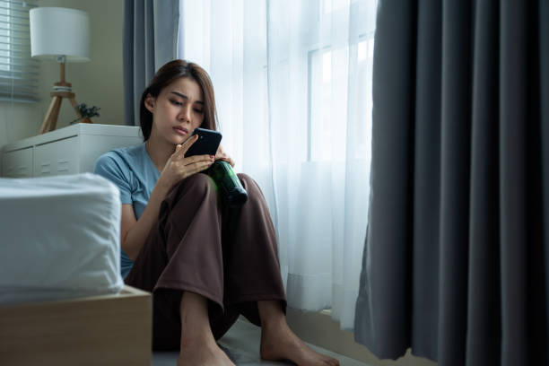 istock Asian young depression woman drinking beer while look at mobile phone. Attractive beautiful girl sitting on floor hold a bottle of alcohol feeling heart broken, drunk and hangover alone in living room 2165308031