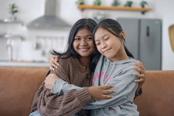 istock Two young girls smiling and hugging while sitting on a couch in a cozy home setting with a modern kitchen in the background 2165314263