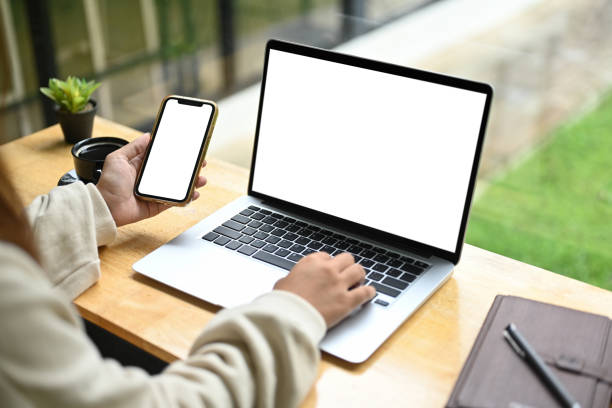Unrecognizable woman holding mobile phone while using laptop on wooden table