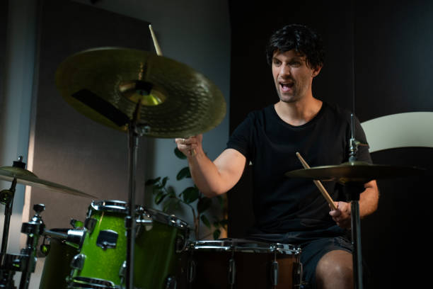 istock A young man sits behind a drum set in a home studio and plays with two drumsticks. The drums are green and black, and there are two cymbals. The drummer is wearing a black t-shirt, and he is looking at the drums as he plays. 2165323035