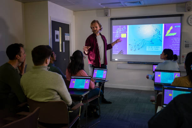 Visual Data Review on Microbiome in the Gut A wide shot of a group of science professionals attending a group seminar is shot from the rear of the audience. The presentation depicts research from gut microbiome data. The audience laptops mirror the main presentation display. A bearded male lecturer is standing at the front of the display with an open palm presenting on-screen data.
Videos are available similar to this scenario. teacher projector stock pictures, royalty-free photos & images