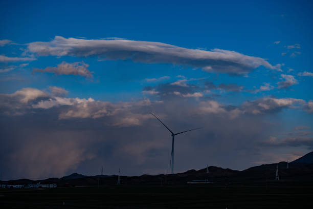 A photo taken at dusk around Sayram Lake, featuring towering wind turbines set against a background of blue sky and clouds. The image captures the contrast between the modern turbines and the natural beauty of the evening sky.