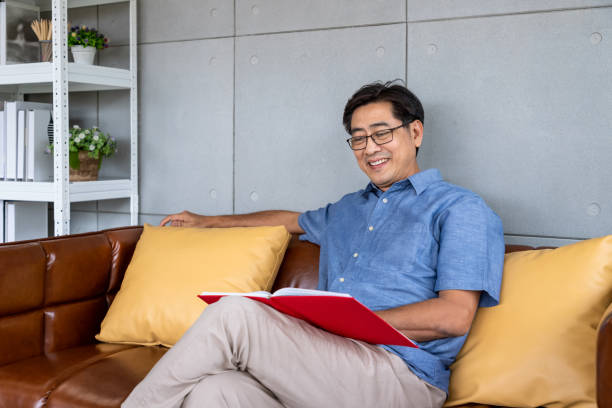 istock Senior Asian man reading book and smiling while relaxing on sofa at home 2165351834