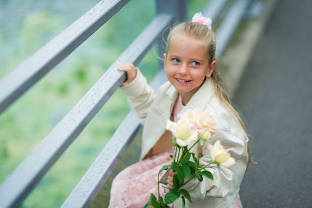 A cheerful young girl with long blonde hair and a pink bow holds a bouquet of flowers while sitting on a bridge. She gazes playfully at the surroundings, enjoying the beauty of spring. The lush greenery in the background complements her bright demeanor and the soft pastel colors of her dress.