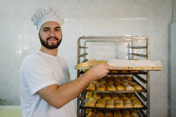 A smiling male baker, dressed in white, holds a tray of freshly baked bread rolls in a bakery with racks full of similar bread in the background.