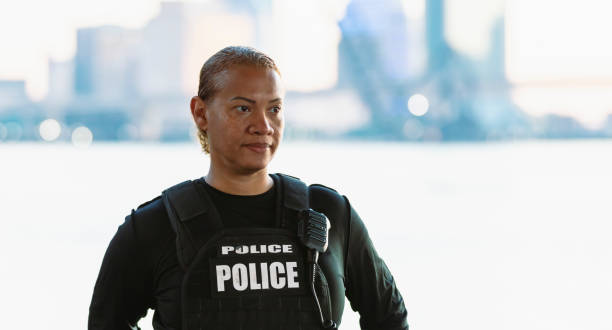 Headshot of a multiracial policewoman standing on a city waterfront, the urban skyline out of focus in the background. The police officer is looking away from the camera with a serious, confident expression. She is a mature woman in her 40s, of African-American, Hispanic and Japanese ethnicity.