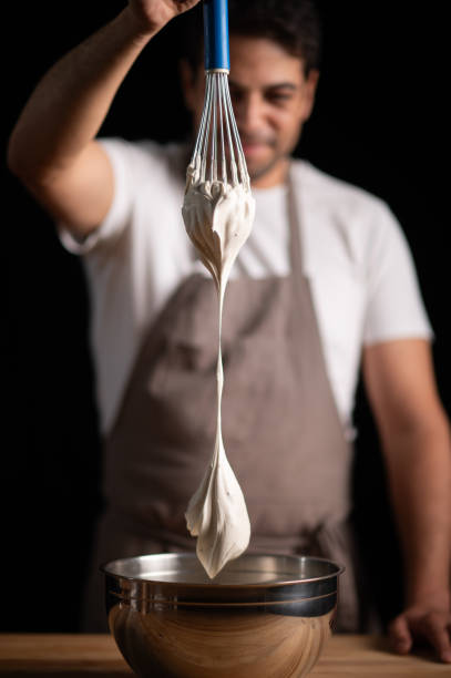 A male pastry chef preparing cream with a wire whisk on a wooden table and dark background.