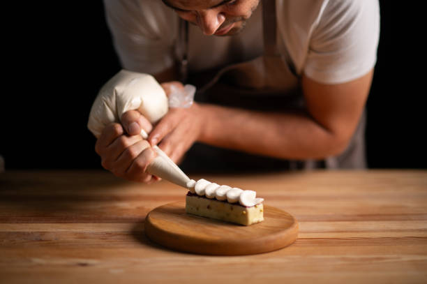 A male pastry chef squeezing cream from icing bag to a cake on a wooden table and dark background.