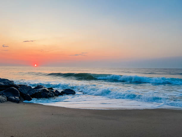 A stunning sunrise paints the sky in vibrant golden, orange, and amber hues above a churning ocean. Gentle waves break onto the sandy shore, creating white foam. A warm, peaceful nature scene perfect for backgrounds.