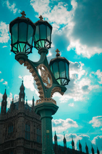 The photograph captures a close-up view of an ornate Victorian-style lamp post in Westminster, with the Palace of Westminster's spires in the background. The lamp post, adorned with intricate gold and green details, stands out against a vibrant blue sky filled with clouds. The architectural elements of the Palace of Westminster are visible, adding a historic and regal context to the image. The combination of the antique lamp post and the Gothic architecture of the palace creates a striking visual contrast, emphasizing the rich heritage of London.