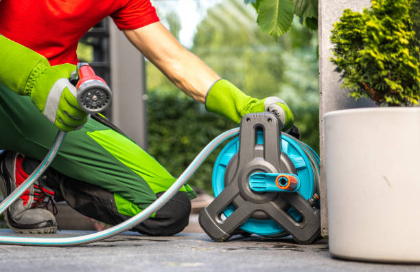 A man in green pants and a red shirt waters plants using a hose and a hose reel. He is kneeling on a patio, holding the nozzle and directing the water towards a plant. The hose is connected to a blue and grey hose reel that is sitting next to the man.