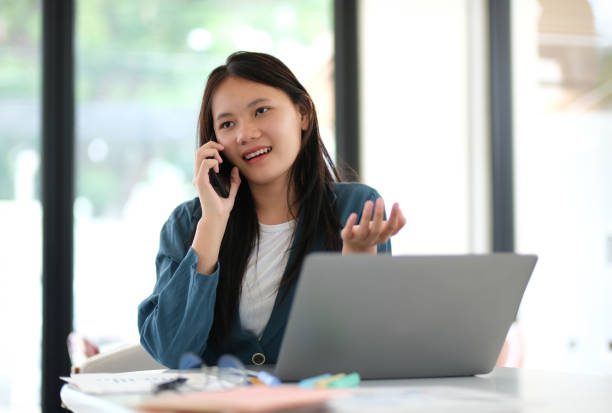 istock A woman is talking on her cell phone while sitting at a desk with a laptop 2165430690