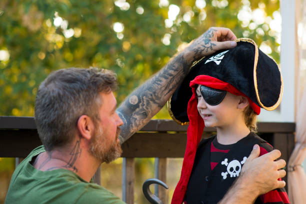 Little elementary age boy is wearing a pirate costume for Halloween trick or treat festivities. His father is helping him put on a big hat with a skull and crossbones on it.