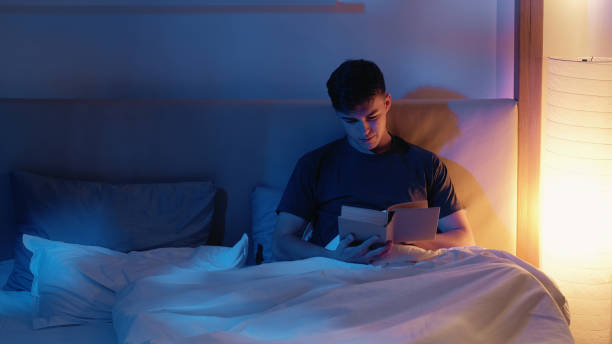 istock Man Reading Book in Cozy Bed With Soft Lighting 2165449807