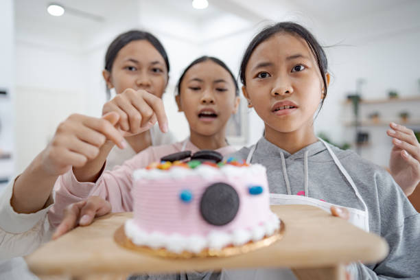 Three young girls in a modern kitchen presenting a decorated cake while filming a cooking tutorial with a smartphone