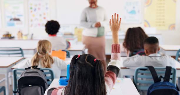 istock Students, classroom and kid with hand raised, questions and knowledge with book, practice and thinking. Answer, school and smart with intelligent, learning and teacher with assignment and education 2165474977