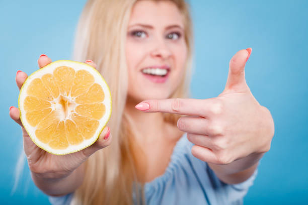Woman smiling girl holding half of green yellow grapefruit citrus fruit in hand, pointing with finger, recommending healthy eating, on blue. Dieting losing weight concept.