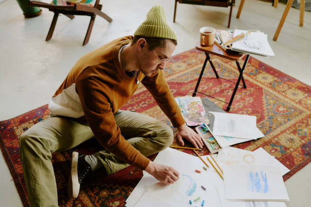 High angle view of young Caucasian man sitting on vintage carpet drawing picture on paper with crayons