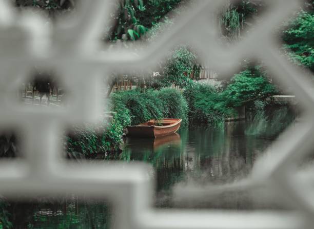 A small wooden boat sits on a tranquil pond, framed by lush greenery, viewed through an ornate white trellis.