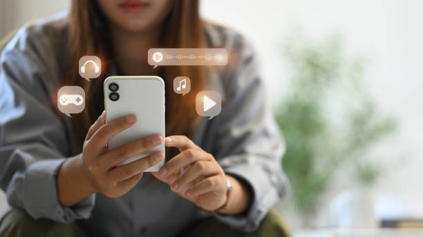 istock Young woman using smartphone, interacting with various multimedia icons including music, video and gaming 2165485128