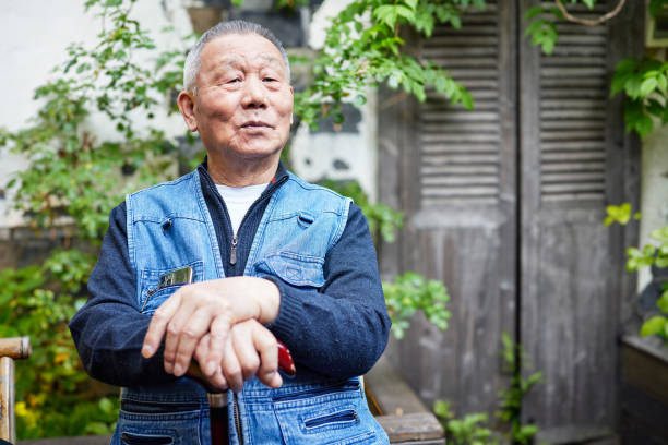 A comfortable spring daytime park with plenty of greenery in Japan. A Japanese man in his late 70s is sitting on a bench wearing a suit.
