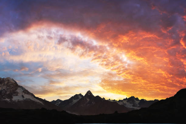 Sun glow and bright orange sunlight in evening cloudy sky behind of incredible mountains with snow and glaciers