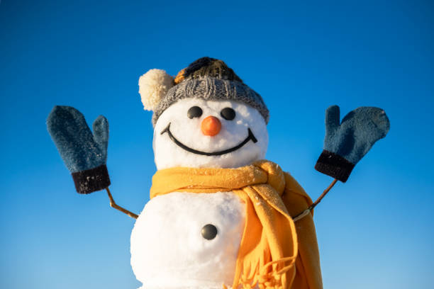 Funny snowman in knitted hat and yellow scalf with hands up on blue sky background. Winter holidays concept