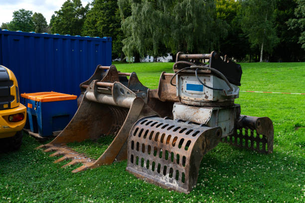 A large mechanical grapple loader sits idle on a green lawn beside a blue shipping container and orange construction equipment under clear skies.