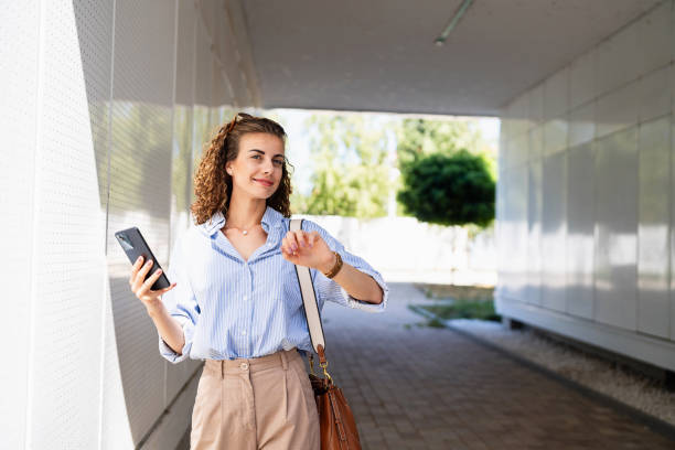 istock Businesswoman using mobile phone and checking the time on her wristwatch while leaving from the office 2165512148