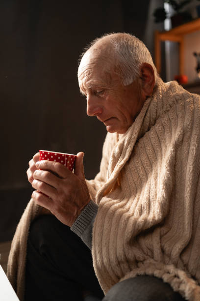 istock Elderly man wrapped in blanket holding a cup of tea, warm home setting 2165515959