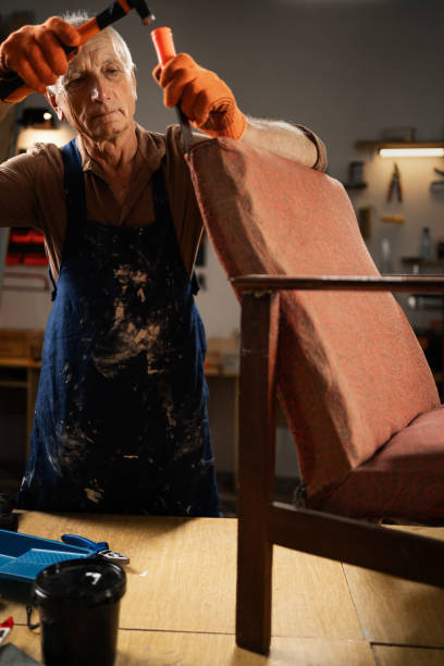 An elderly man in gloves and a blue apron begins to restore an old chair in a workshop. holds a hammer removes old fabric. concept second life of old furniture. Copy space. Place for text.