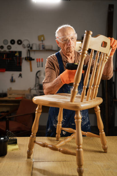 istock An elderly man in a workshop uses a drill with a special attachment to remove old varnish from a wooden chair. He is wearing protective glasses and orange gloves. 2165516132