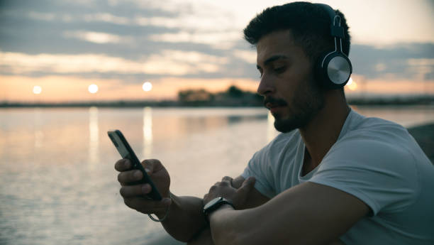 istock Young man in headphones sitting on beach at sunset using smartphone 2165518704