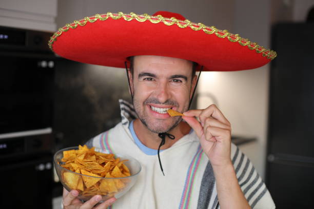 A handsome man in his early forties is eating some delicious nachos. He wears a poncho and a Mexican sombrero and holds a large bowl with corn snacks.
