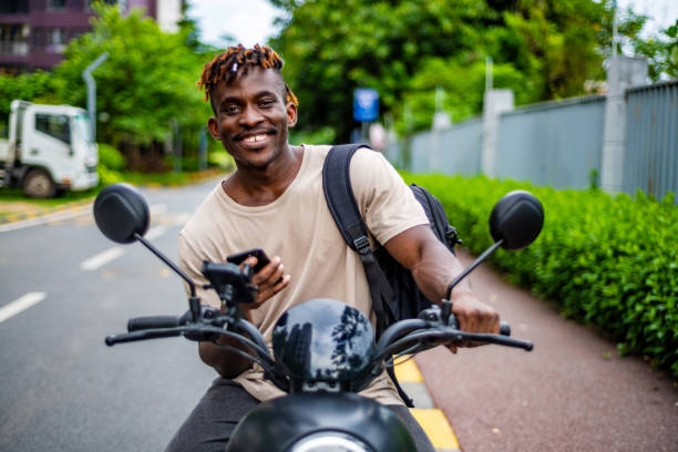 A cheerful black man with a stylish hairstyle sits on a motorbike, engaged with his smartphone, wearing casual attire in an urban setting.