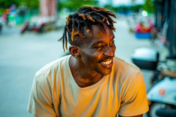 A smiling Black man with trendy dreadlocks sits outside, dressed in casual attire, radiating happiness in an urban setting.