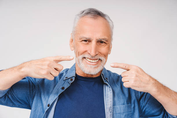 Happy mature gray-haired man pointing with hands at teeth looking at camera over white background