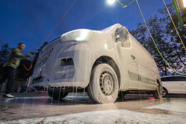 A worker applies thick foam to a van at a car wash in the evening. The vehicle, partially covered in foam, is illuminated by overhead lights, creating a vibrant atmosphere as the day transitions to night.