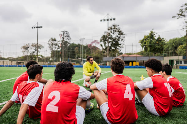 A group of young men sits on the grass at a soccer field in Sydney, Australia, taking a break from training. They are engaged in light conversation and enjoying their time together.