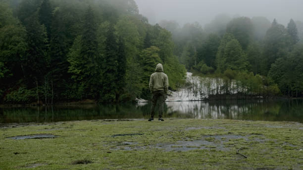 Man stands facing a serene, misty lake surrounded by lush forest. Solitude, nature, and tranquility, themes of reflection and outdoor adventure.
