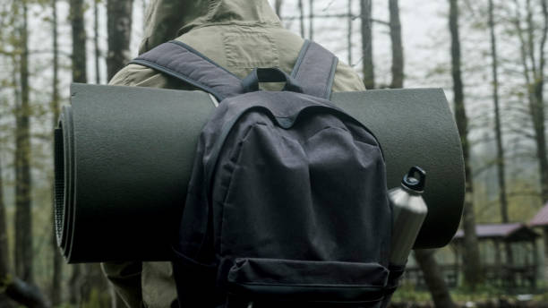 A close-up of a hiker's backpack in a forest. The backpack is equipped with a rolled-up sleeping mat and a metal water bottle, indicating preparation for a camping or trekking adventure.